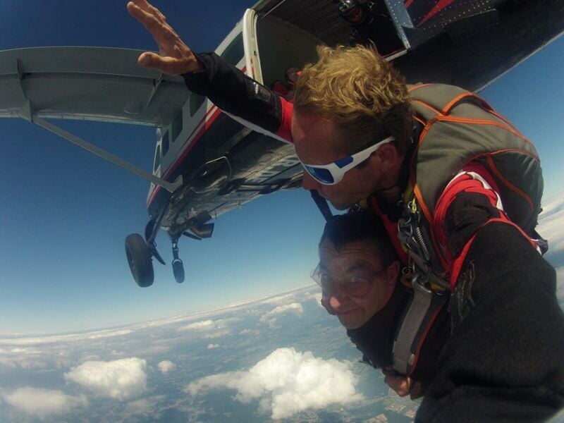 Deux personnes en tandem sautent en parachute depuis un avion, ciel bleu et nuages en arrière-plan.
