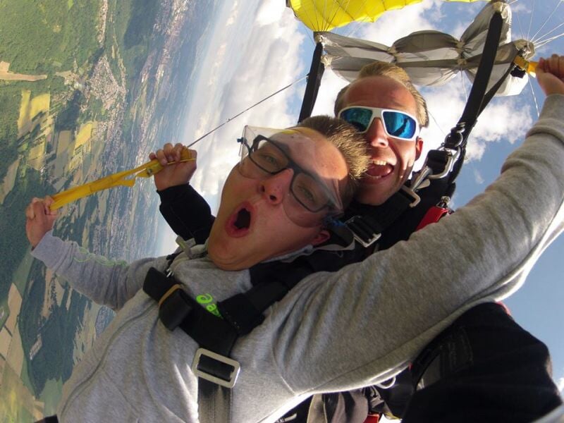 Deux hommes en tandem en parachute, l'un fait une expression de surprise, ciel et paysage en dessous.
