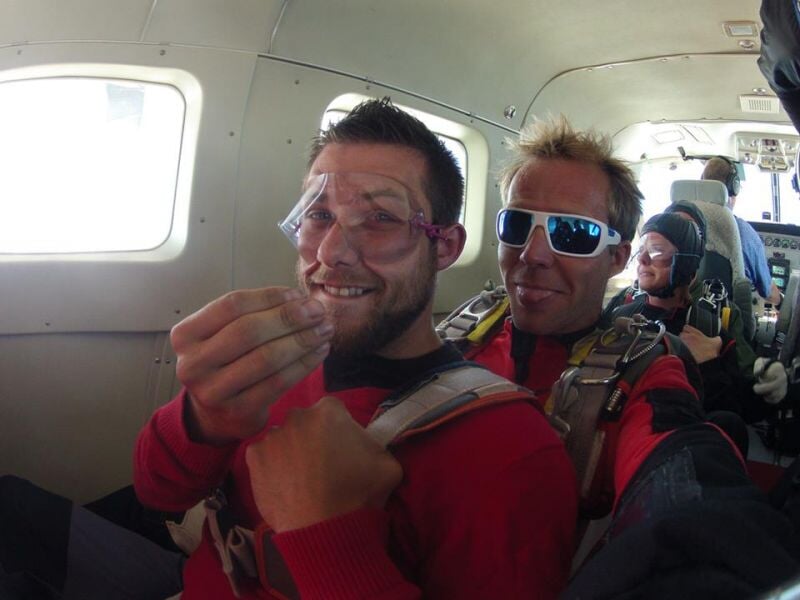 Deux hommes souriants à l'intérieur d'un avion avant un saut en parachute, équipement de sécurité visible.