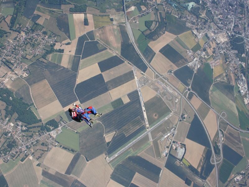 Vue aérienne d'un parachutiste en tandem au-dessus de champs et routes, paysage rural étendu.
