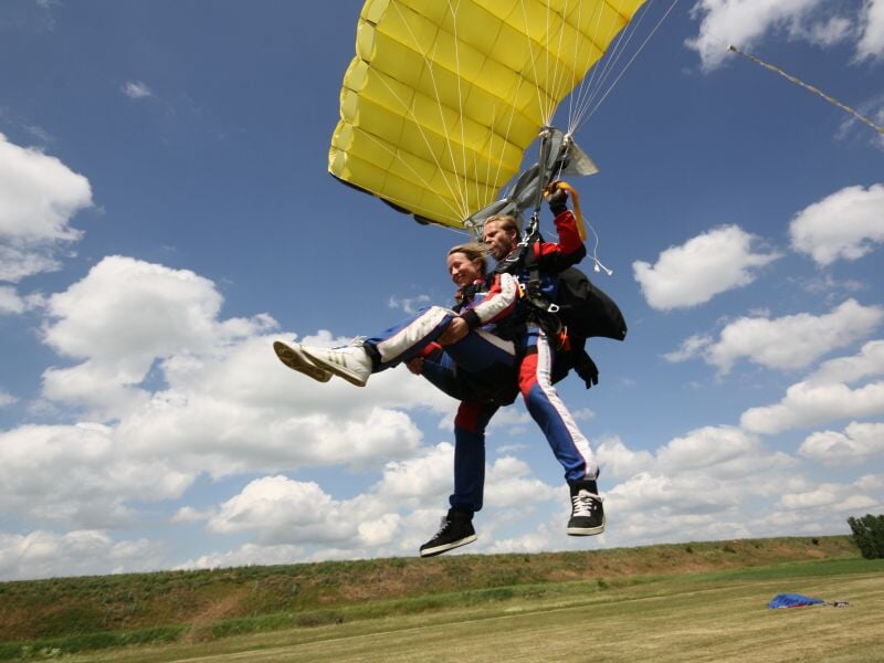 Deux parachutistes en tandem atterrissant sur un terrain herbeux sous un ciel partiellement nuageux.