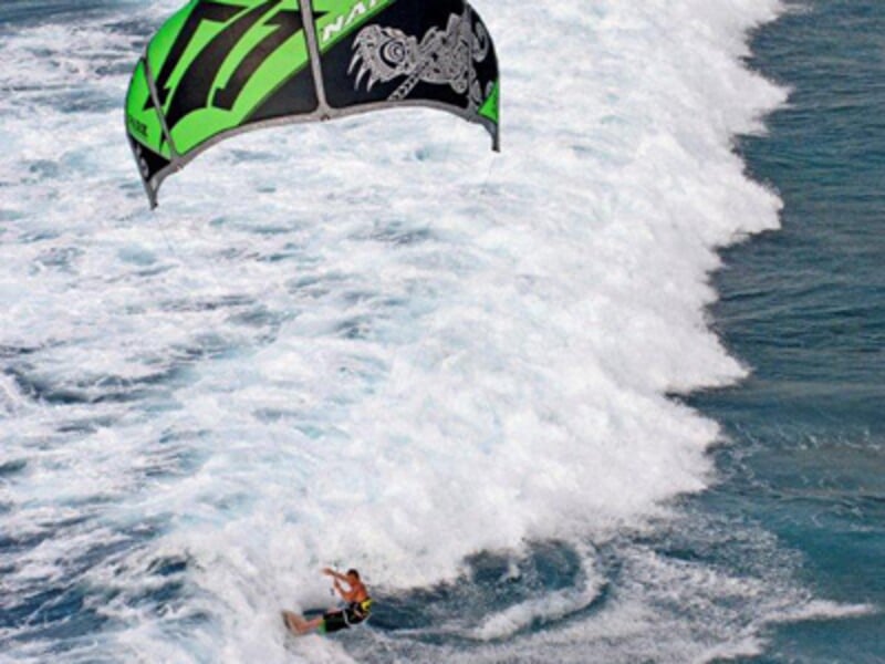 Kitesurfeur glissant sur une vague blanche avec une grande voile verte et noire au-dessus de l'eau agitée.