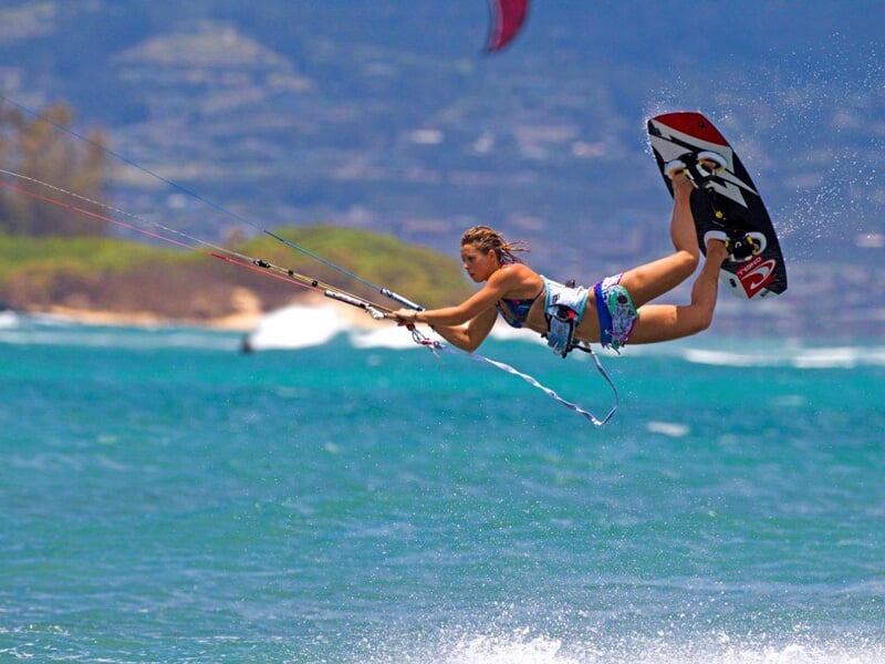 Femme kitesurfeuse en vol au-dessus de l'eau turquoise, tenant la barre de contrôle avec un paysage montagneux en arrière-plan.