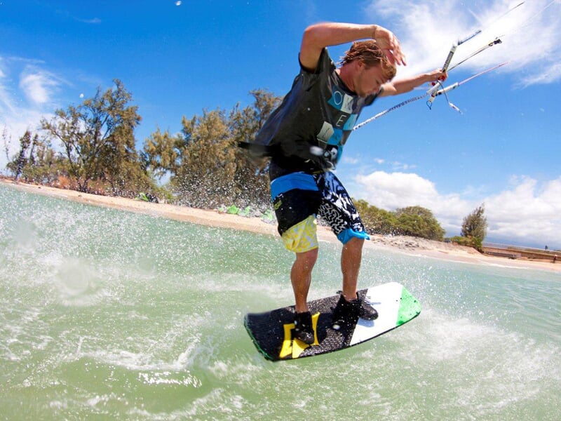 Homme kitesurfeur en équilibre sur sa planche verte, créant des éclaboussures près du rivage avec des arbres en arrière-plan.