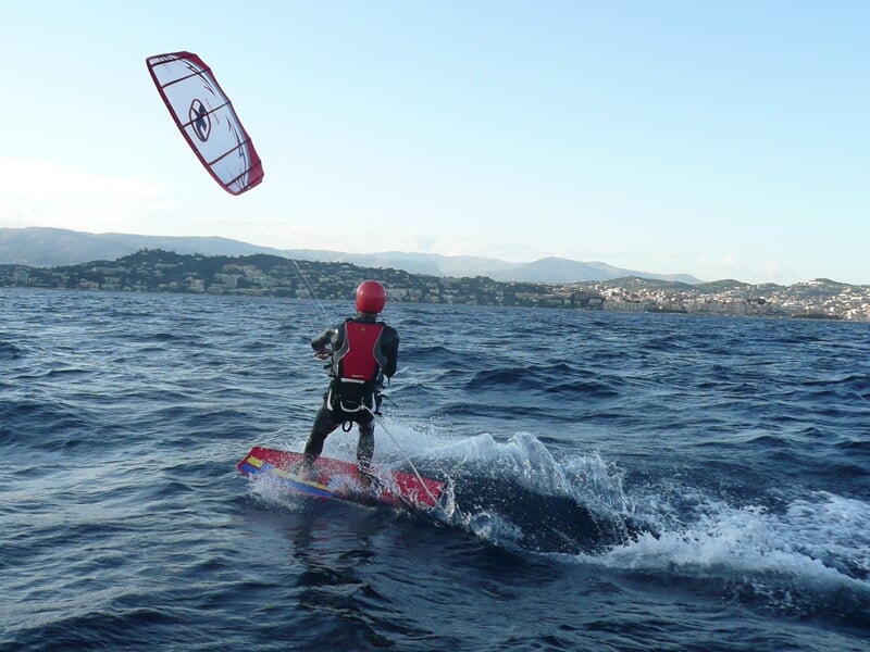 Kitesurfeur vu de dos sur une mer agitée, portant un casque rouge et un gilet de sécurité, avec des collines au loin.