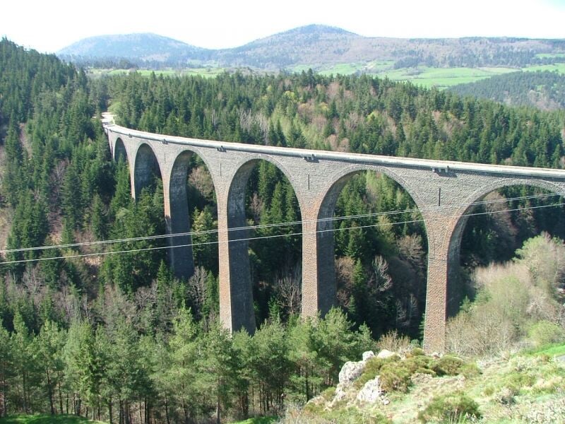 Vue panoramique d'un viaduc en pierre à plusieurs arches traversant une forêt dense, avec des collines au loin sous un ciel bleu clair