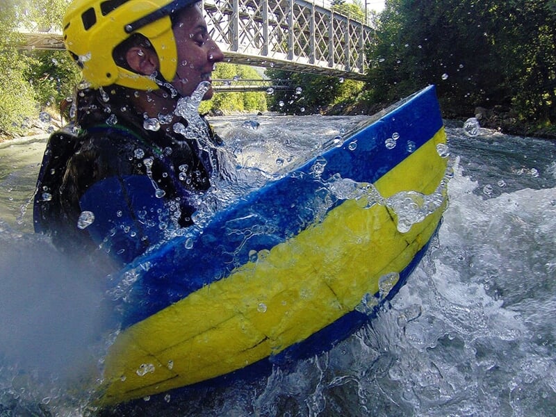 Personne en hydrospeed avec casque jaune naviguant dans les rapides d'une rivière sous un pont métallique.