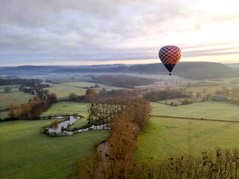 Montgolfière colorée survolant une campagne avec un ruisseau sinueux et des arbres en contrebas.