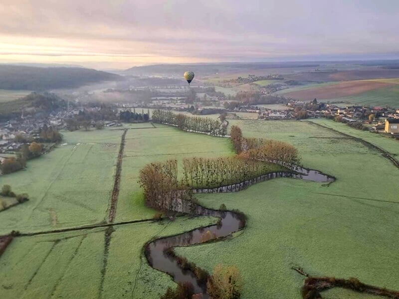 Vue aérienne d’une vallée verdoyante avec un ruisseau sinueux et une montgolfière au loin.