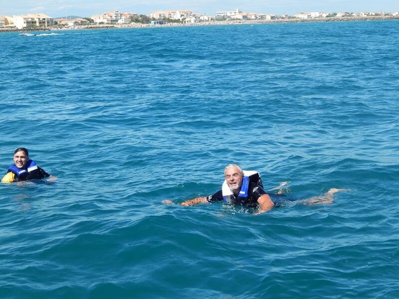 Deux personnes nageant dans une mer bleue claire, portant des gilets de sauvetage, sous un ciel dégagé.