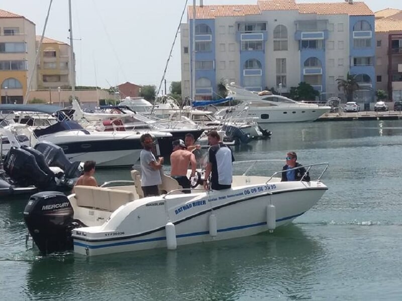 Un petit bateau blanc avec plusieurs personnes à bord naviguant dans un port avec des immeubles en arrière-plan.