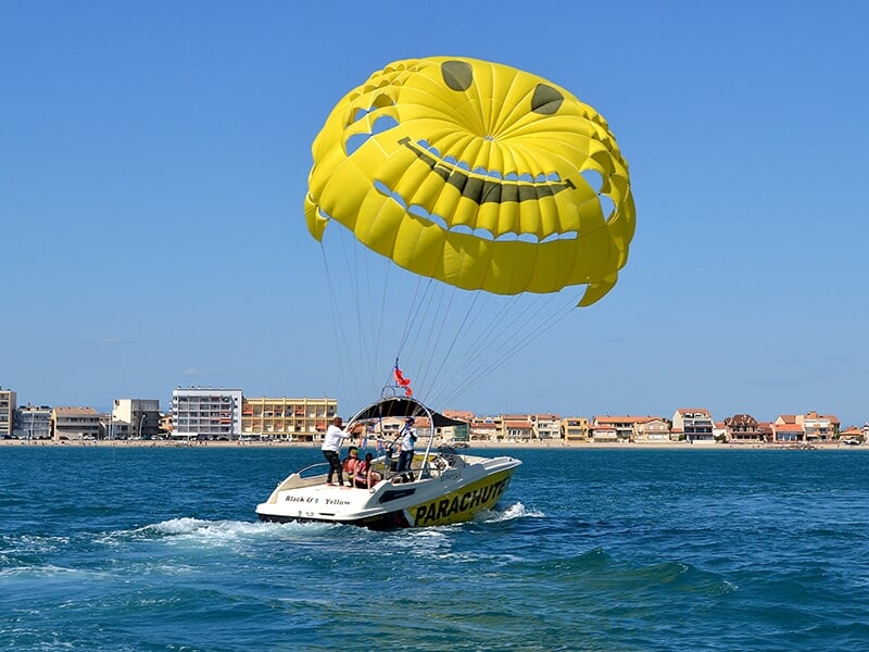 Un bateau blanc tractant un parachute jaune souriant avec plusieurs personnes à bord naviguant près du rivage.