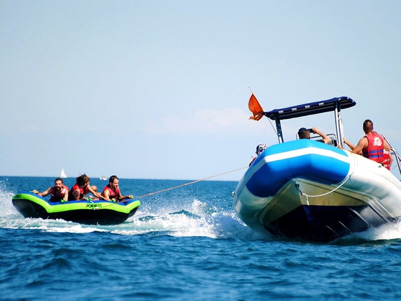 Un bateau bleu tracte une bouée verte et noire avec plusieurs personnes à bord sur une mer bleue sous un ciel clair.
