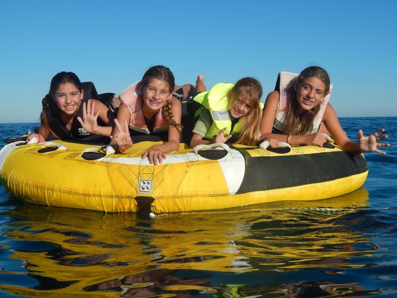 Quatre enfants souriants allongés sur une bouée jaune tractée sur une mer calme sous un ciel bleu clair.