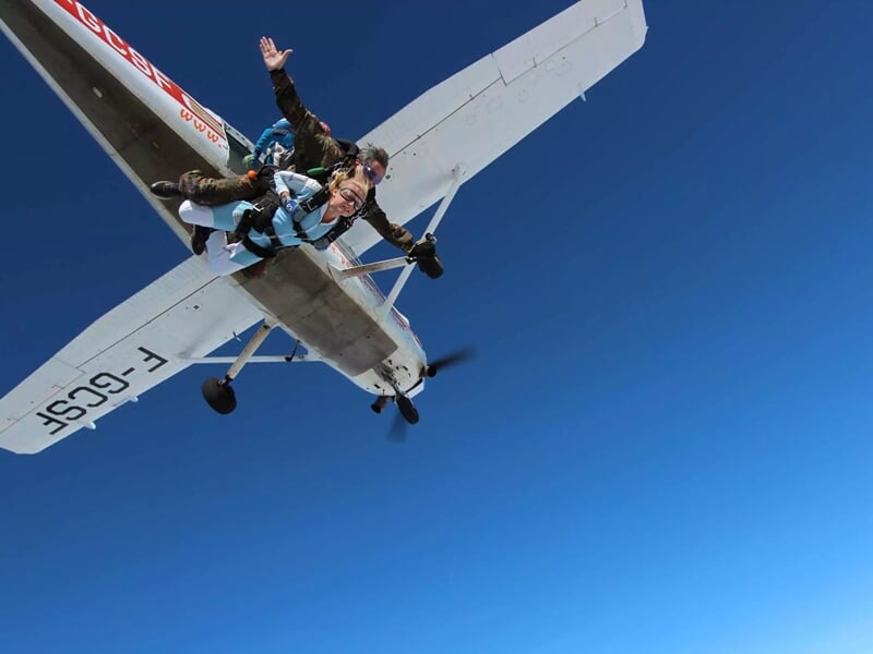 Deux personnes effectuent un saut en parachute depuis un petit avion blanc sous un ciel bleu clair sans nuages.