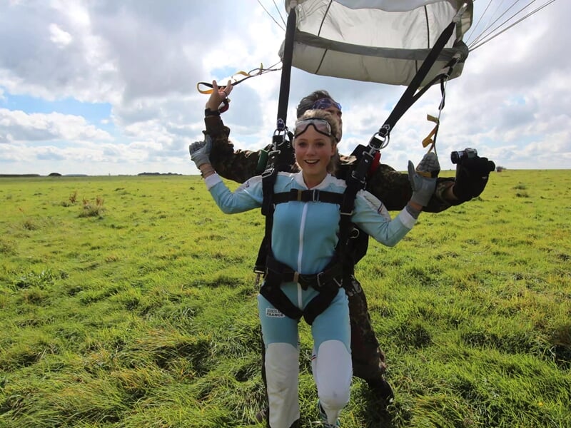 Une femme souriante en combinaison bleue atterrit en parachute sur un champ vert avec un instructeur derrière elle.