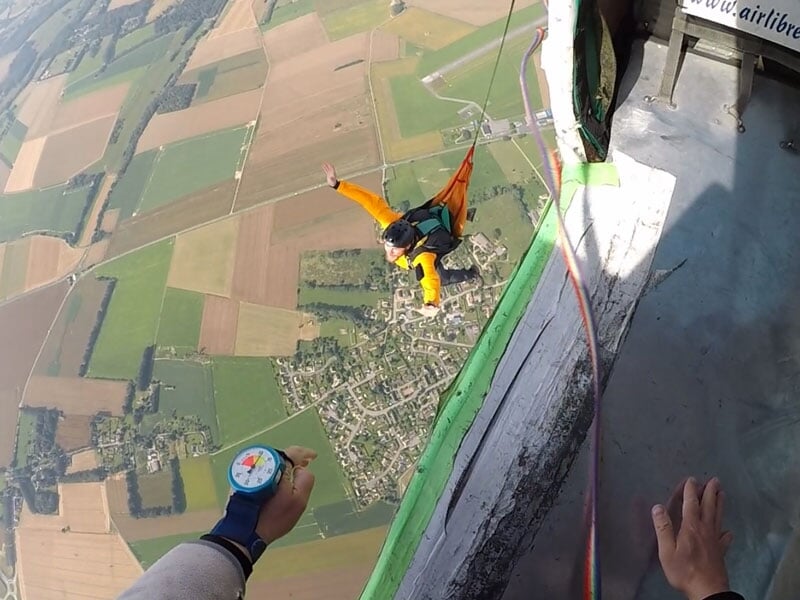 Saut en parachute d'un participant en veste orange et casque noir, suspendu à une corde attachée à une plateforme. Vue aérienne sur des champs et un village en contrebas, avec un altimètre coloré visi