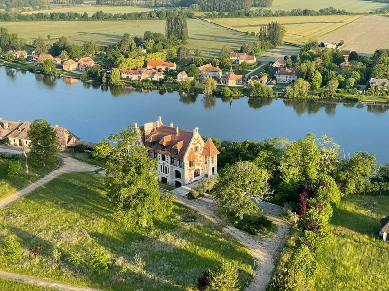 Maison ancienne au bord d'une rivière calme, entourée d'arbres et d'espaces verts ensoleillés.
