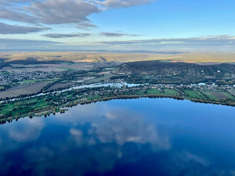 Vue aérienne d'un lac entouré de collines et de zones habitées sous un ciel bleu avec quelques nuages.