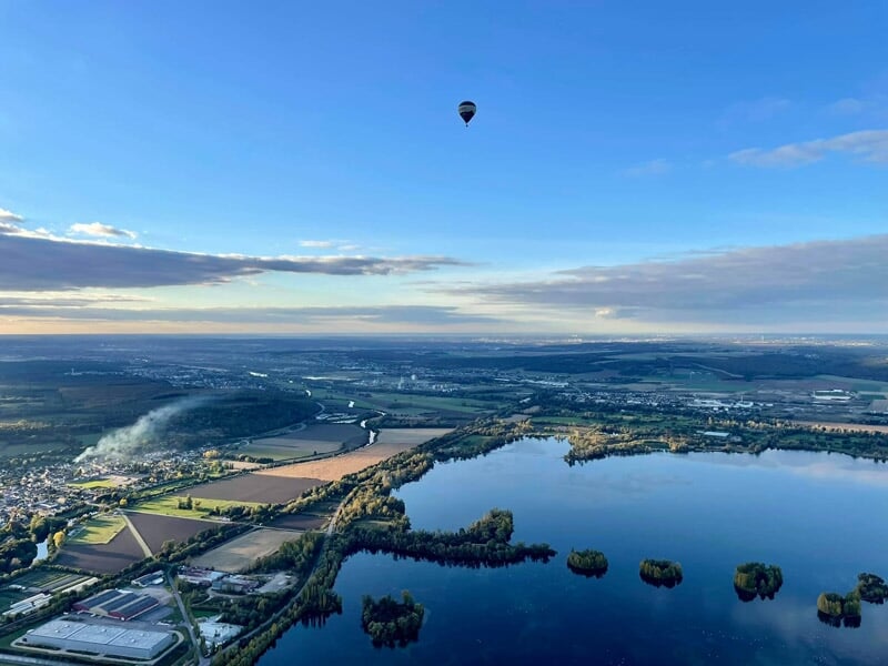 Montgolfière noire et bleue flottant au-dessus d'un lac et d'un paysage rural sous un ciel clair.