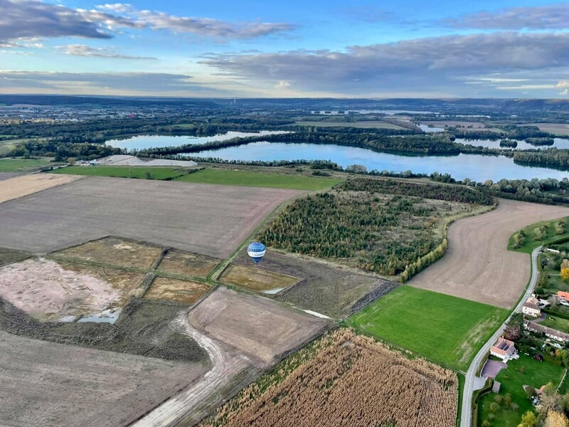 Montgolfière bleue et blanche avec inscription, survolant un champ vert et un lac en arrière-plan.