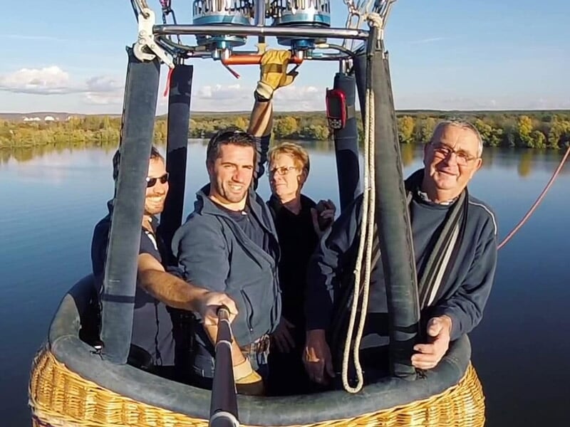 Montgolfière colorée en vol au-dessus d'un paysage rural avec champs, forêts et plans d'eau.