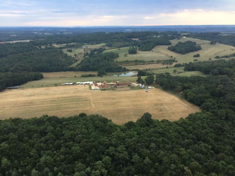 Paysage rural vu du ciel avec champs cultivés, bâtiments agricoles et forêt dense en arrière-plan.
