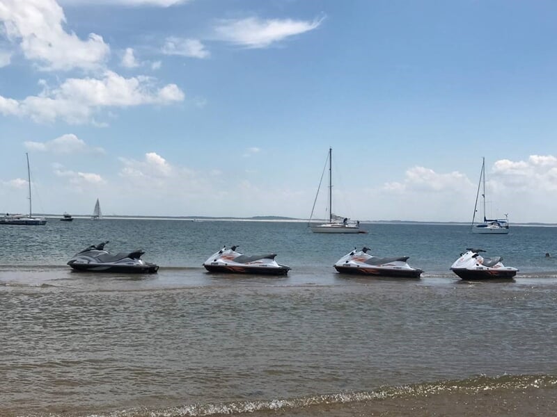 Quatre jets skis alignés sur une plage de sable avec des voiliers au loin sur une mer calme.