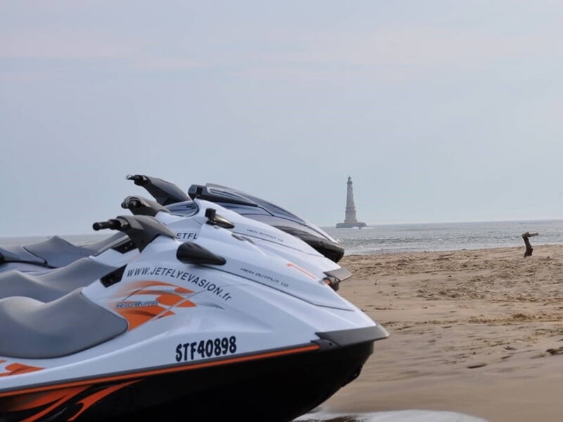Plusieurs jets skis stationnés sur une plage de sable avec un phare visible au loin sur la mer.
