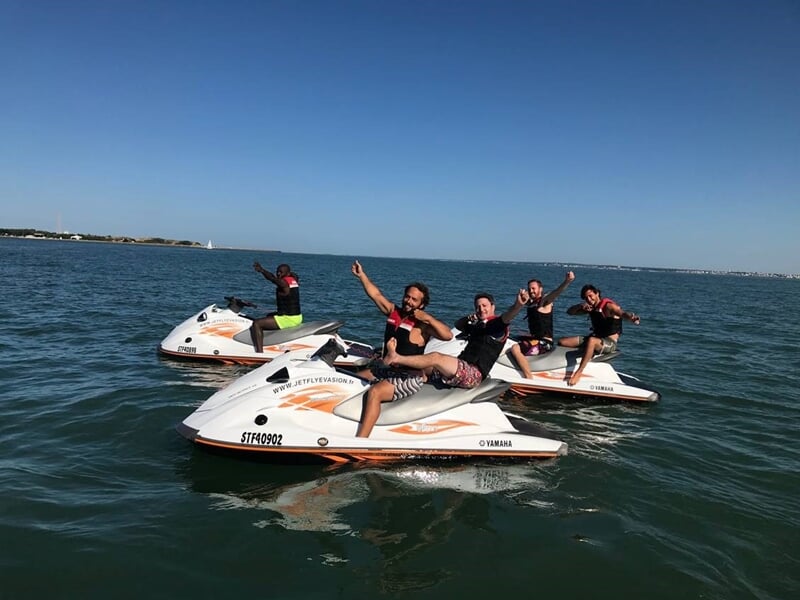 Quatre personnes souriantes sur des jets skis en mer, saluant la caméra sous un ciel dégagé.
