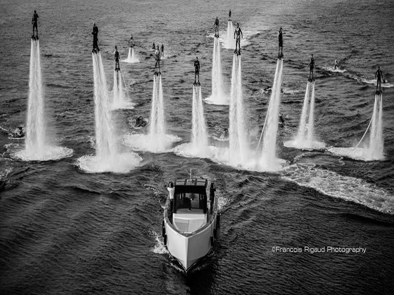 Groupe de dix personnes en flyboard s'élèvent en formation au-dessus de la mer, derrière un bateau.