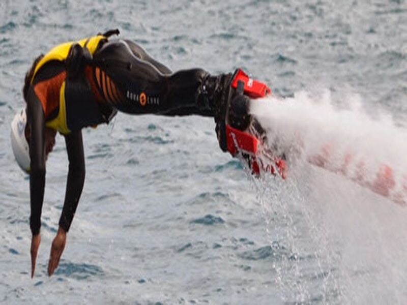 Personne en combinaison de protection effectue un plongeon acrobatique avec un flyboard propulsé par jets d'eau.