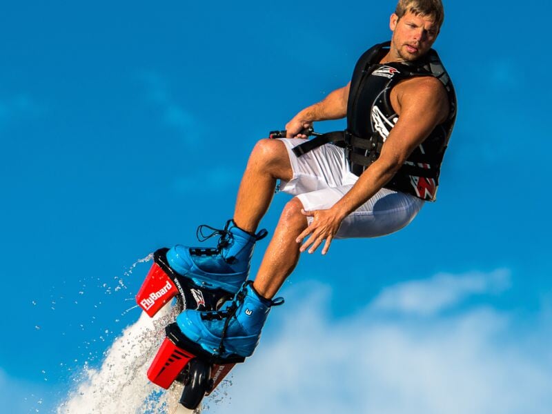 Homme en flyboard avec gilet de sauvetage bleu, en plein vol au-dessus de l'eau sous un ciel bleu clair.