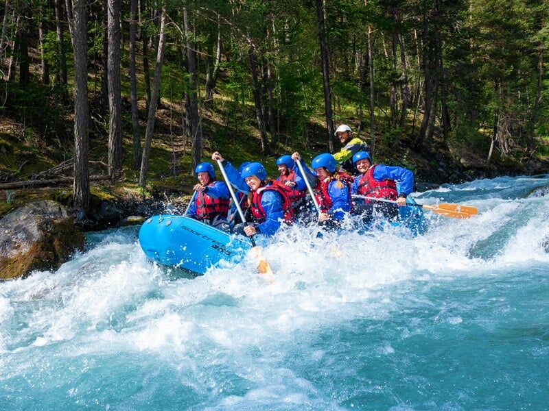 Groupe de six personnes en tenue de rafting pagayant dans un raft bleu sur une rivière en montagne.