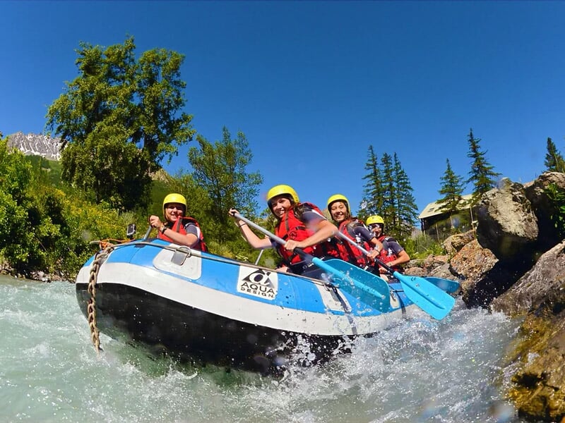 Quatre personnes en casque jaune pagayant dans un raft bleu sur une rivière avec des arbres en arrière-plan.