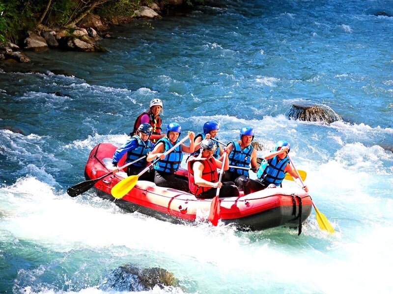 Groupe de six personnes en gilets de sauvetage pagayant dans un raft rouge sur une rivière agitée.
