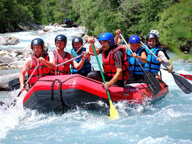 Six personnes en raft rouge pagayant avec énergie dans une rivière de montagne entourée de verdure.