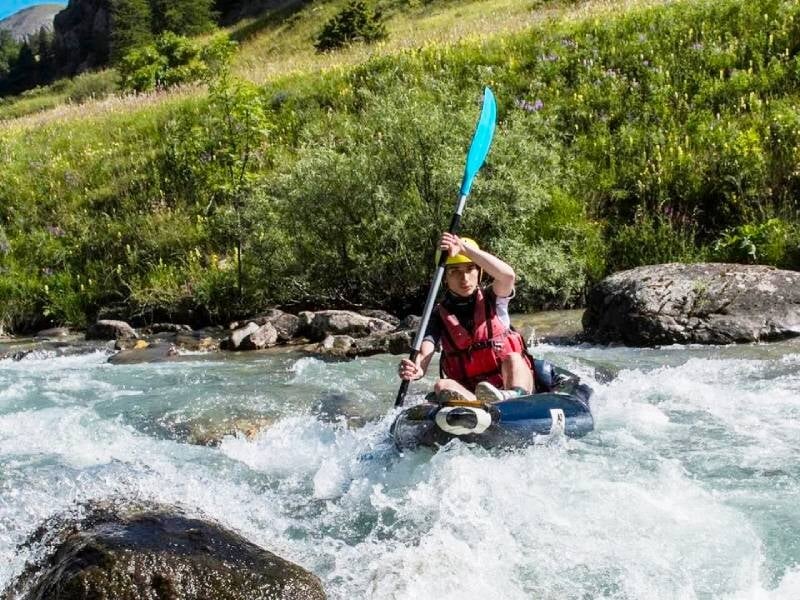 Personne en casque rouge pagayant dans un kayak bleu sur une rivière de montagne avec des rapides.