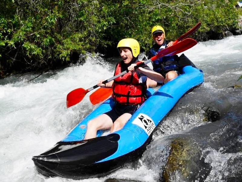 Deux personnes souriantes en gilets rouges pagayant dans un kayak bleu sur une rivière agitée.