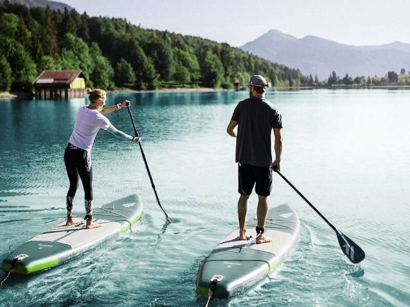 Deux personnes debout sur des planches de stand-up paddle sur un lac calme entouré de montagnes.