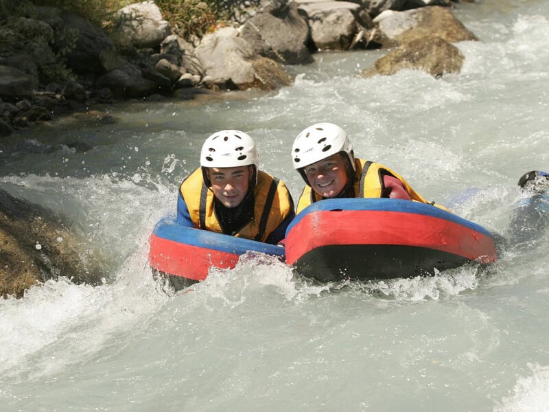 Deux personnes souriantes en casque blanc sur des flotteurs rouges dans une rivière agitée.