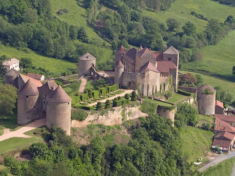 Vue aérienne d'un château médiéval entouré de verdure et de petits bâtiments dans un paysage vallonné.