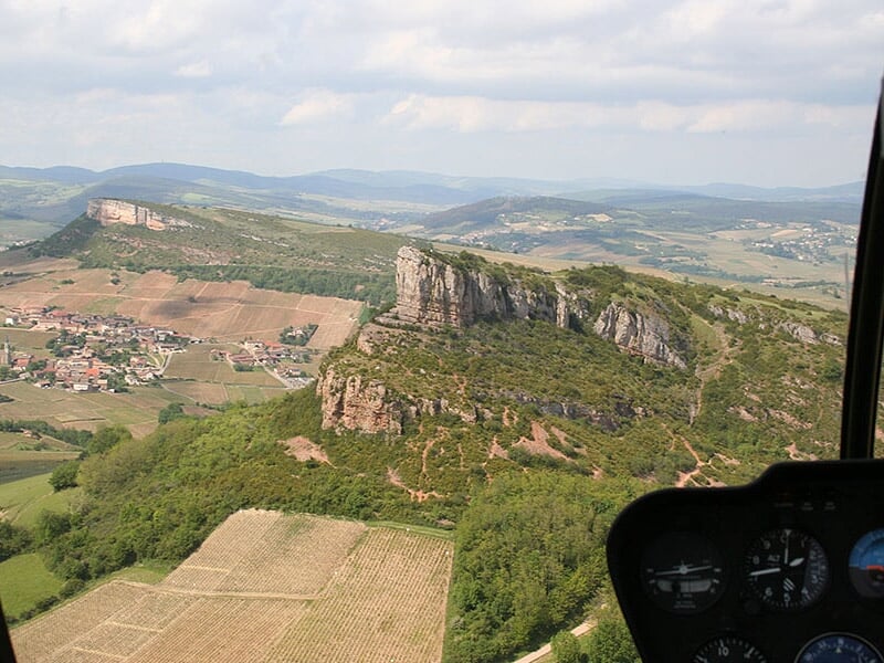 Vue depuis un hélicoptère montrant une formation rocheuse imposante au milieu de collines et de vignobles.