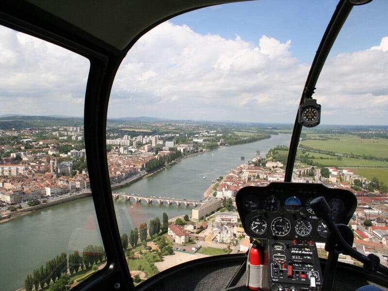 Cockpit d'hélicoptère avec vue sur une ville traversée par une rivière et un pont, sous un ciel partiellement nuageux.