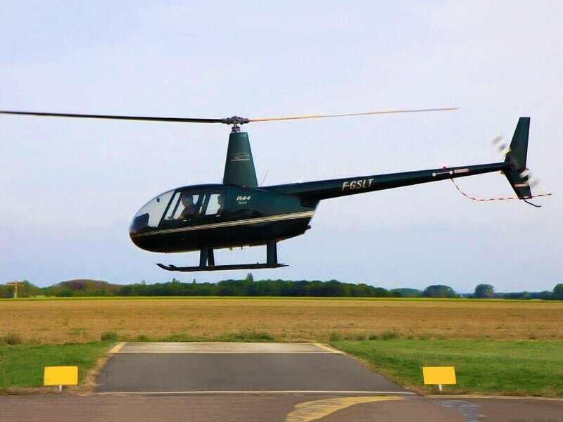 Hélicoptère noir en vol stationnaire au-dessus d'une piste d'atterrissage dans un champ vert.