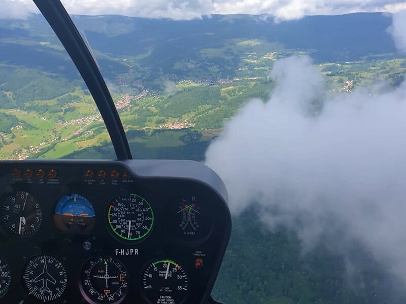 Tableau de bord d'un hélicoptère en vol avec vue sur des collines et des nuages à l'extérieur.