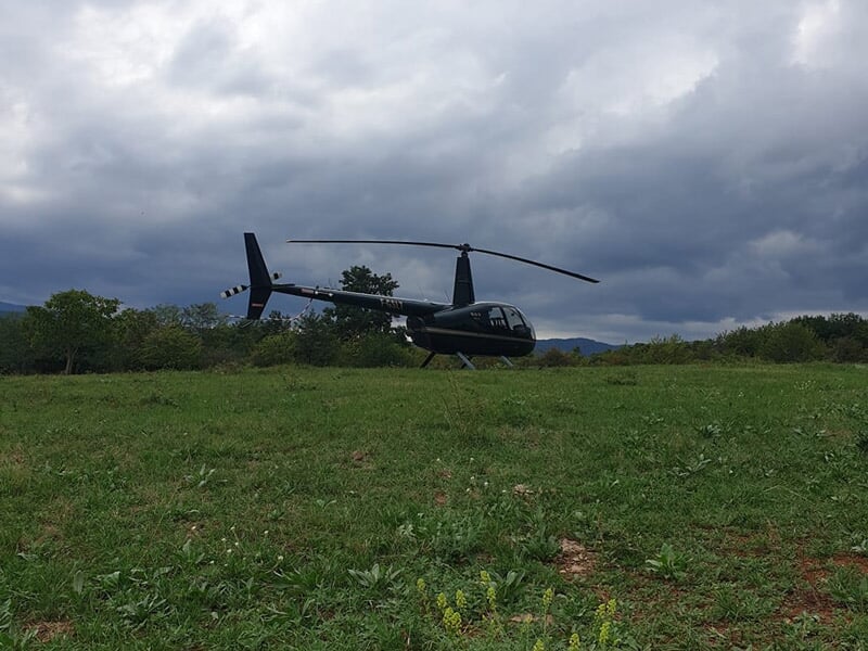 Hélicoptère noir posé sur un terrain herbeux sous un ciel nuageux avec des arbres en arrière-plan.