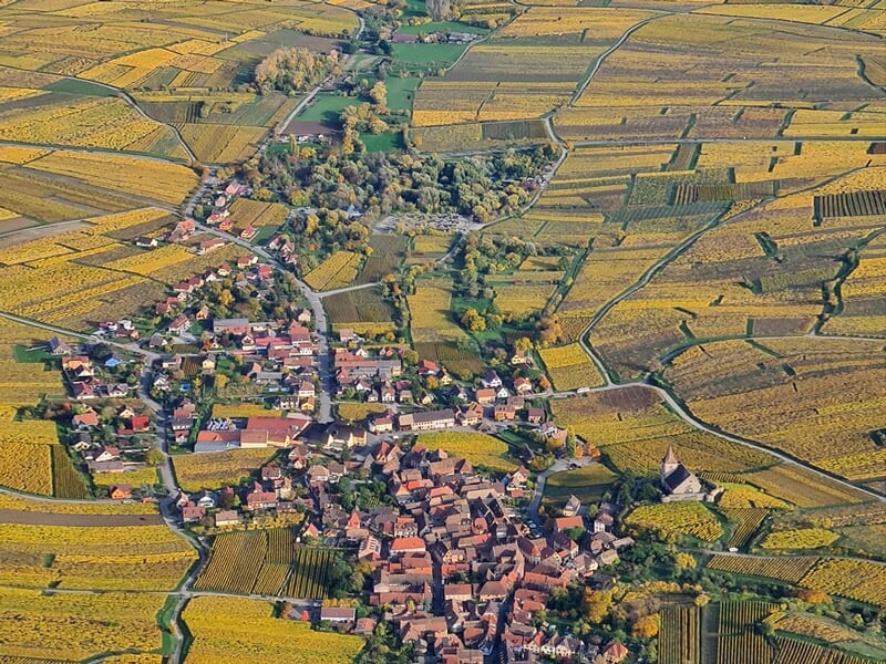 Vue aérienne d'un village entouré de vastes champs jaunes et verts sous un ciel partiellement nuageux.