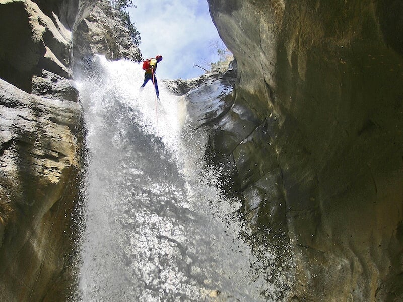 Personne en combinaison descend une cascade en rappel au milieu d’une gorge rocheuse, l’eau éclaboussant autour de lui.