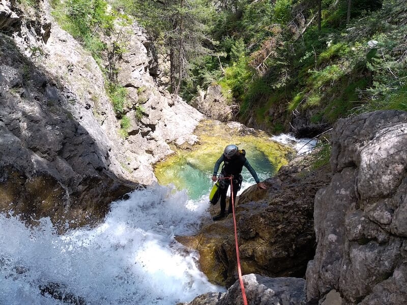 Personne équipée de cordes et casque progressant le long d’une cascade au cœur d’un canyon verdoyant.